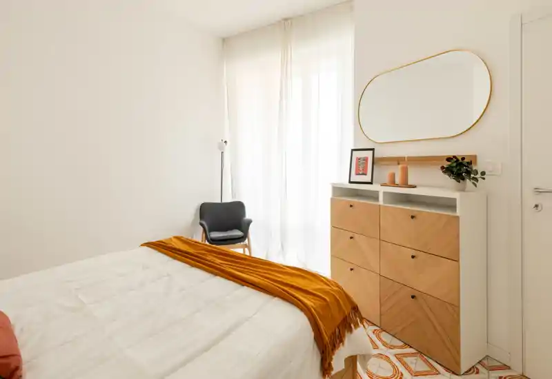 Chambre lumineuse au style doux avec un lit double en linge blanc et un plaid ocre chaleureux. La lumière naturelle traverse des voilages; une commode en bois et un fauteuil apportent une atmosphère minimaliste et accueillante.