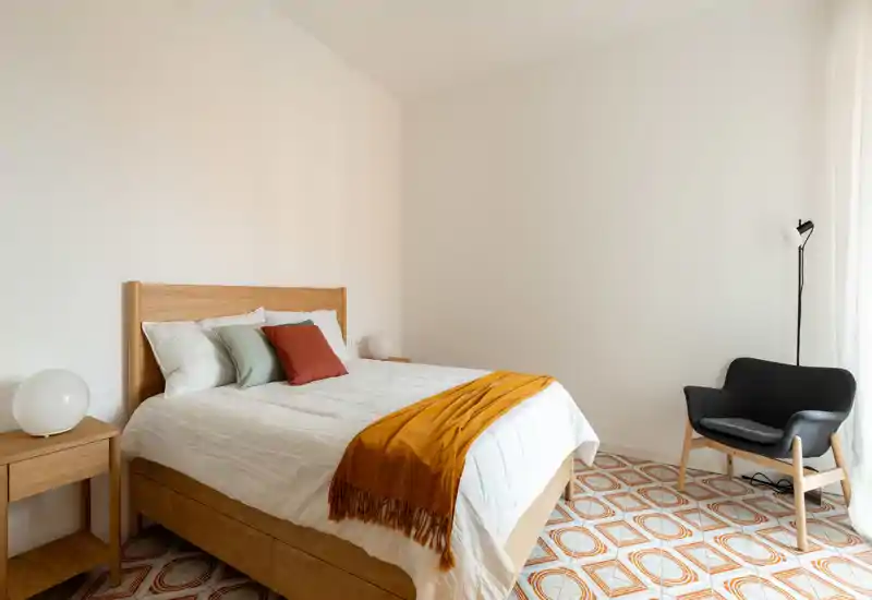 Chambre lumineuse et minimaliste avec cadre de lit en bois, linge de lit blanc, coussins colorés et un plaid ocre chaleureux. La lumière naturelle et le sol décoratif créent un espace accueillant et soigné, parfait pour une annonce.