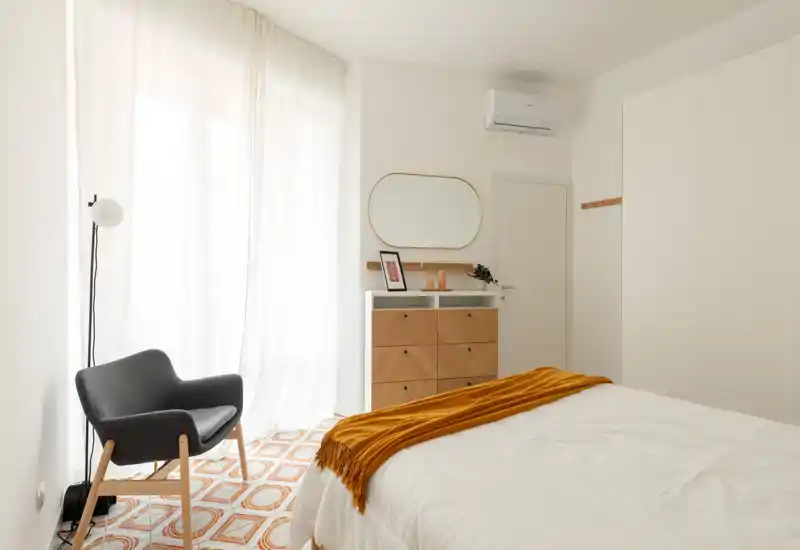 Chambre lumineuse et minimaliste avec un lit en linge blanc et un plaid ocre, un fauteuil moderne près de rideaux légers et une commode en bois avec miroir — espace aéré et bien mis en scène pour une annonce.