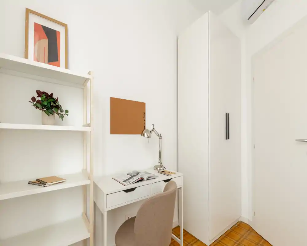 Coin de chambre lumineux et compact avec bureau blanc, chaise et armoire ; étagère minimaliste et tableau en liège forment un espace de travail soigné.