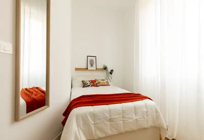 Chambre lumineuse et minimaliste avec un lit simple confortable, coussins décoratifs et un plaid rouge chaleureux ; la lumière naturelle tamisée par des voilages crée une ambiance cosy.