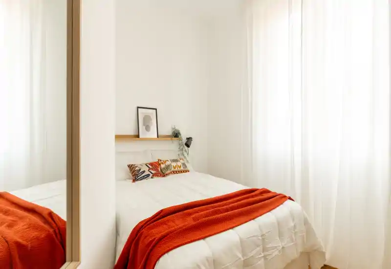 Chambre lumineuse et chaleureuse avec un lit bien fait, coussins décoratifs et un plaid orange; la lumière naturelle filtrée par les voilages crée une ambiance calme et accueillante.