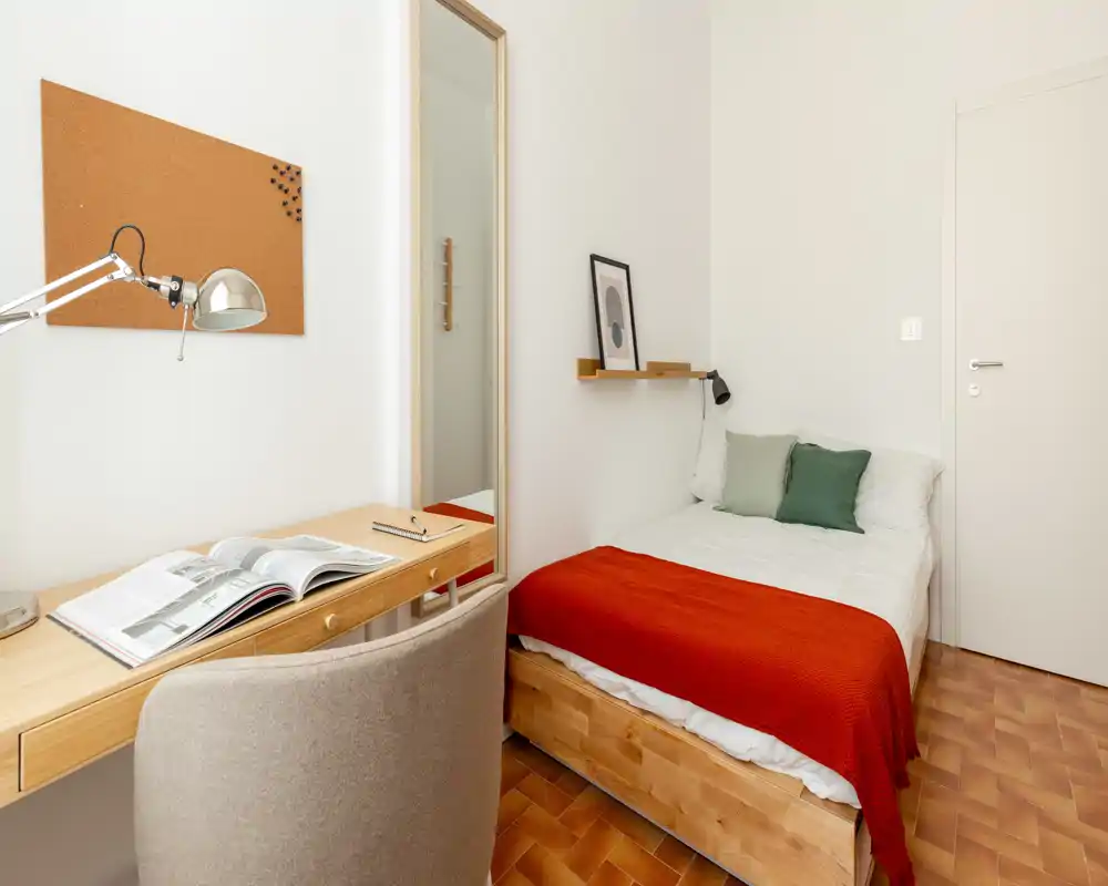 Bright, well-styled single bedroom with a wooden bed frame, crisp white linens and a red throw; includes a compact desk and mirror for a functional study corner.