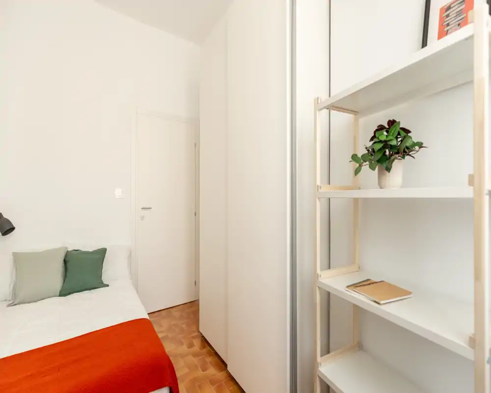 Compact, bright bedroom with a neatly made single bed, green cushions and an orange throw. A tall wardrobe and open shelving provide practical storage in a minimalist, well-lit layout.