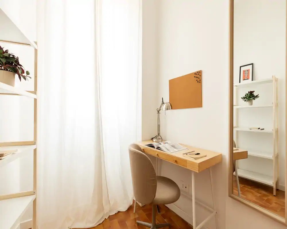 Bright, minimal home office corner with a wooden desk, comfortable swivel chair, corkboard and shelving — ideal for focused work in a cozy, sunlit space.