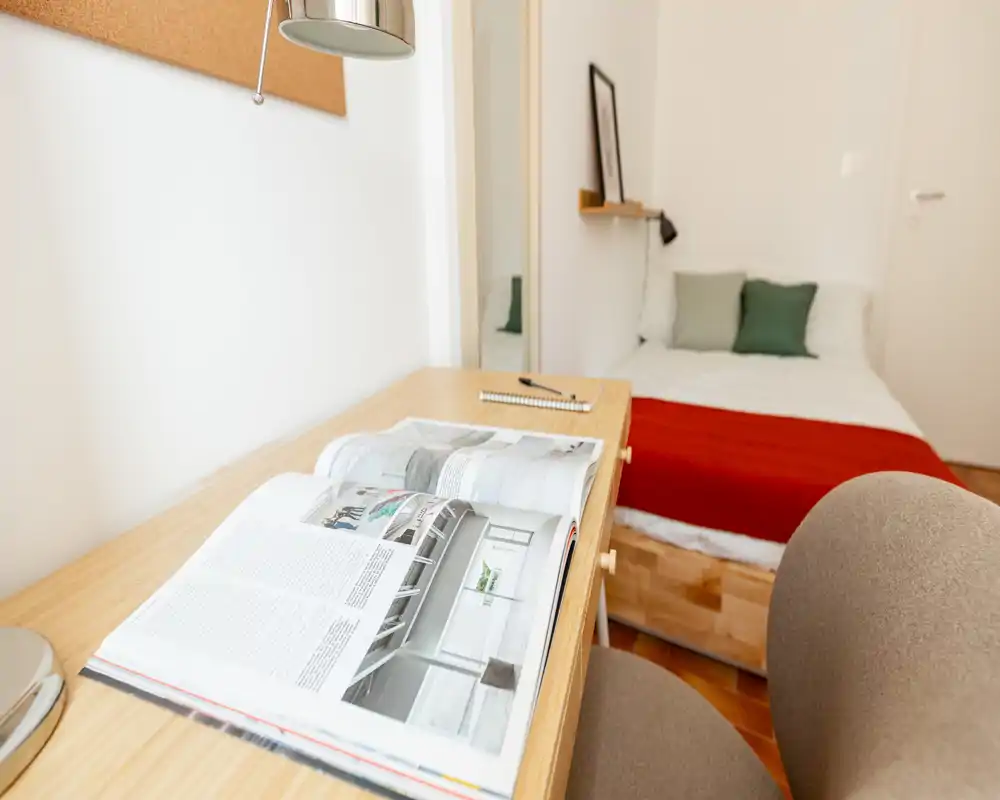 Bright, tidy single bedroom with a wooden bed frame, white linens and a red throw. A small desk with an open magazine and a chair occupies the foreground, creating a cozy study corner.