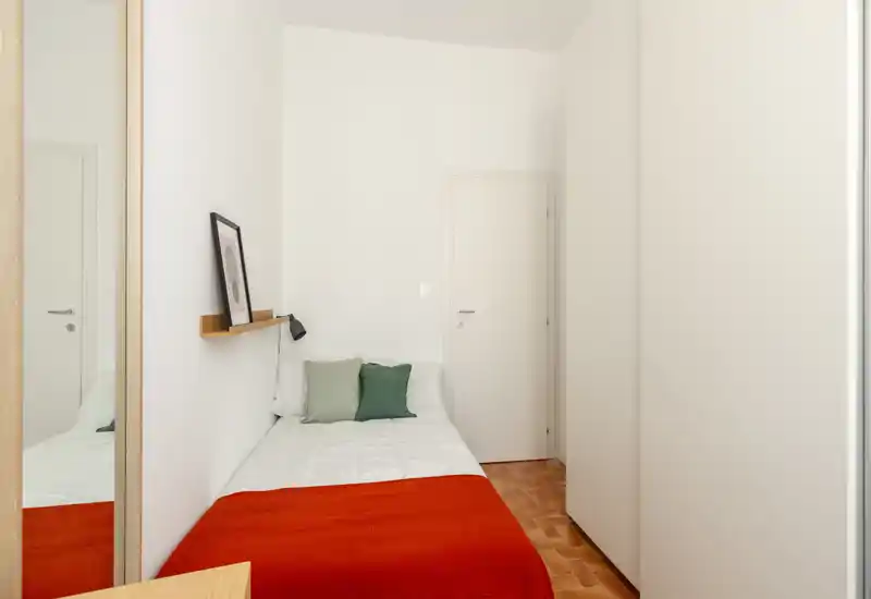 Chambre compacte et lumineuse avec un lit simple en linge blanc et couvre-lit rouge, deux coussins décoratifs, une étagère murale avec un cadre et une lampe — simple, cosy et bien éclairée.
