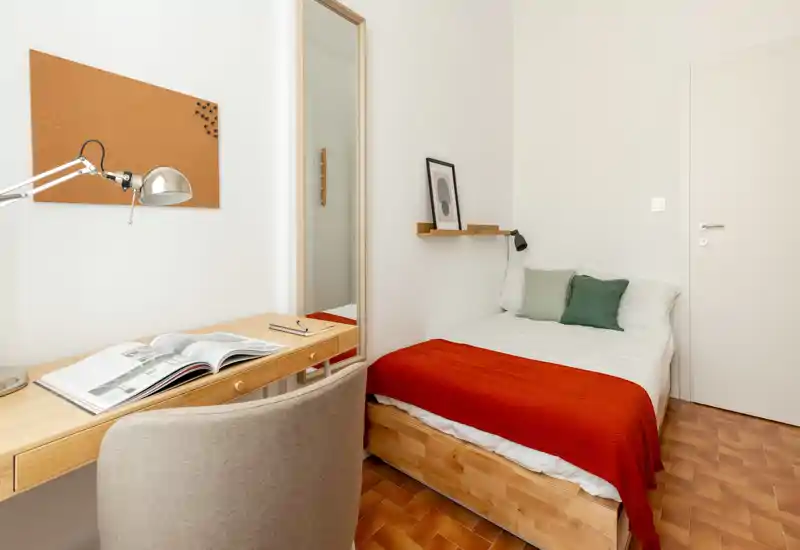 Chambre simple lumineuse et bien décorée avec lit en bois, linge de lit blanc et jeté rouge ; petit bureau et miroir pour un coin travail fonctionnel.