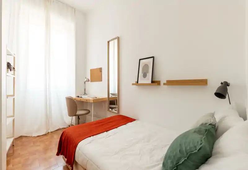 Chambre lumineuse et minimaliste avec un lit confortable, coussins verts et jeté orange; la lumière naturelle entre par une grande fenêtre et un petit bureau offre un espace de travail.