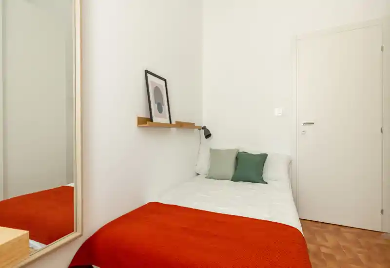 Petite chambre simple et lumineuse avec lit bien fait, coussins verts et un plaid rouge chaleureux. Décor moderne et épuré, étagère murale, lampe de lecture et armoire miroir pour agrandir l’espace.