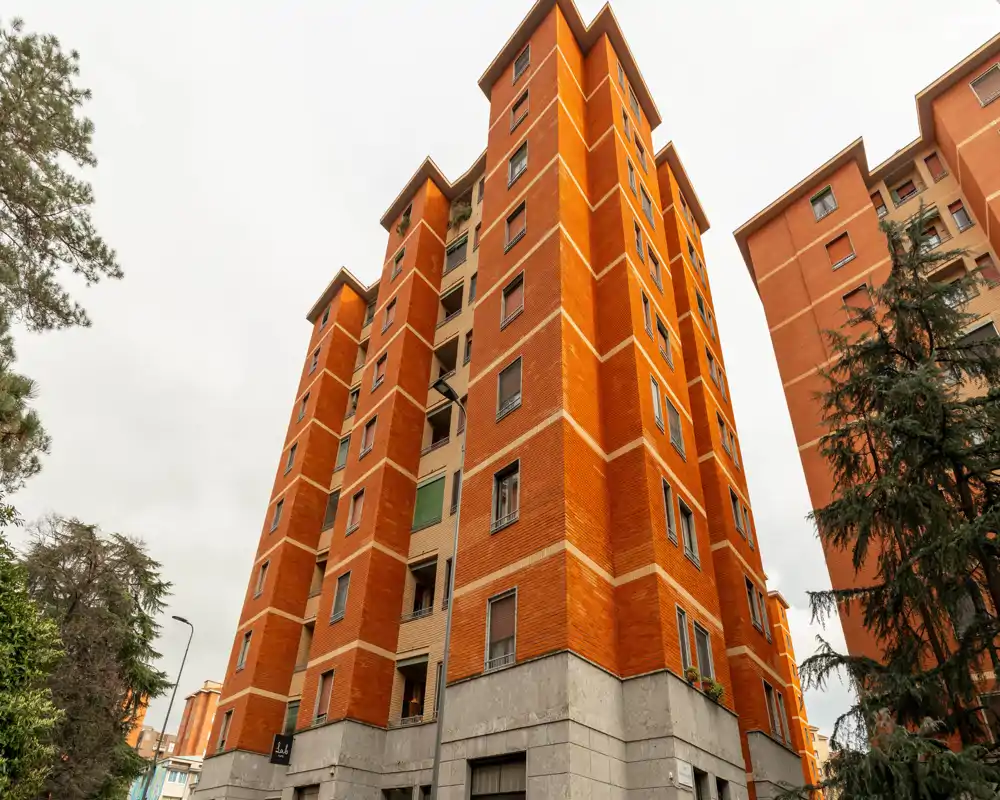 Vue extérieure d'un immeuble résidentiel en brique de plusieurs étages avec des arbres et des bâtiments voisins, adaptée pour présenter la façade et l'emplacement.