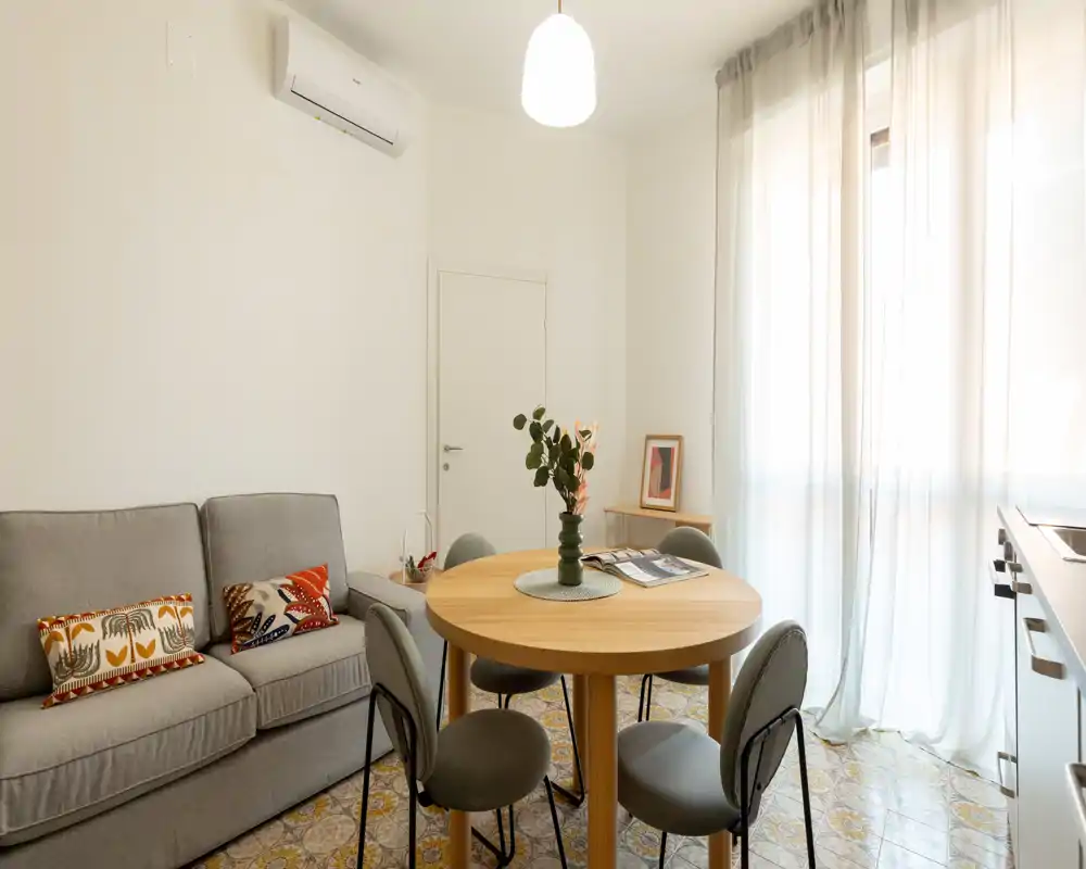 Coin séjour lumineux et élégant avec un canapé gris confortable, une table ronde en bois et quatre chaises. Grande fenêtre avec voilages diffusant une douce lumière naturelle; coussins décoratifs et vase apportent de la chaleur.