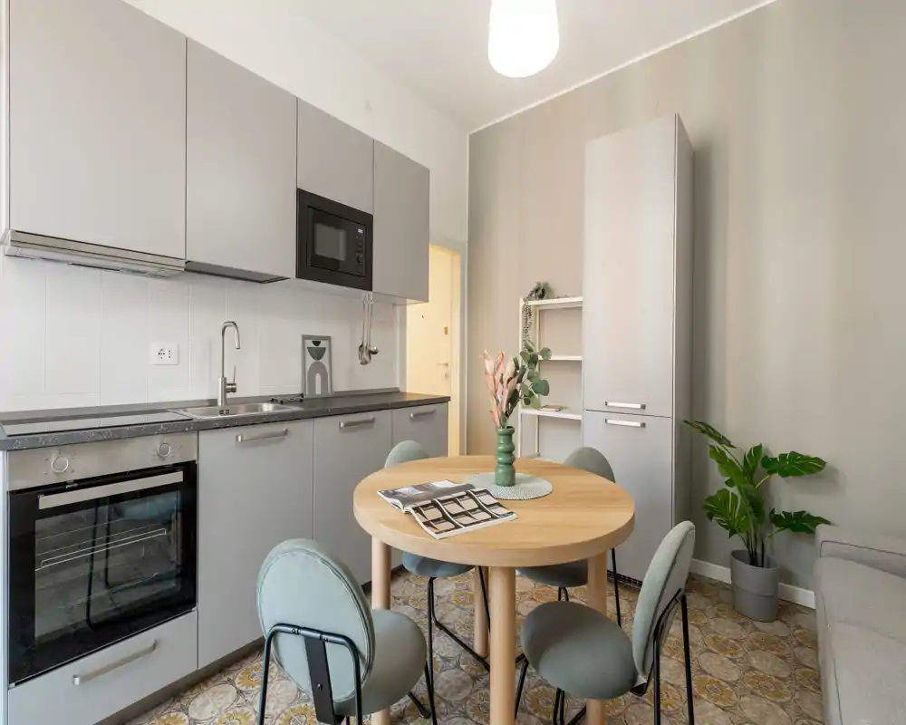 Cuisine moderne et compacte avec placards gris, four et micro-ondes intégrés, et une table ronde en bois avec quatre chaises — lumineuse, stylée et bien organisée pour un petit appartement.