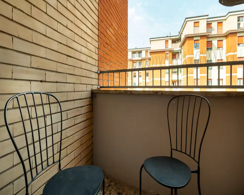 Petit balcon urbain avec deux chaises en métal, mur en briques et vue sur des immeubles voisins ; lumière du jour agréable et coin extérieur simple et fonctionnel.