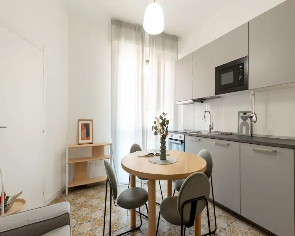 Cuisine compacte et moderne avec rangements intégrés et une table ronde. La lumière naturelle traverse des voilages légers; les meubles gris et le bois apportent une atmosphère chaleureuse et fonctionnelle.