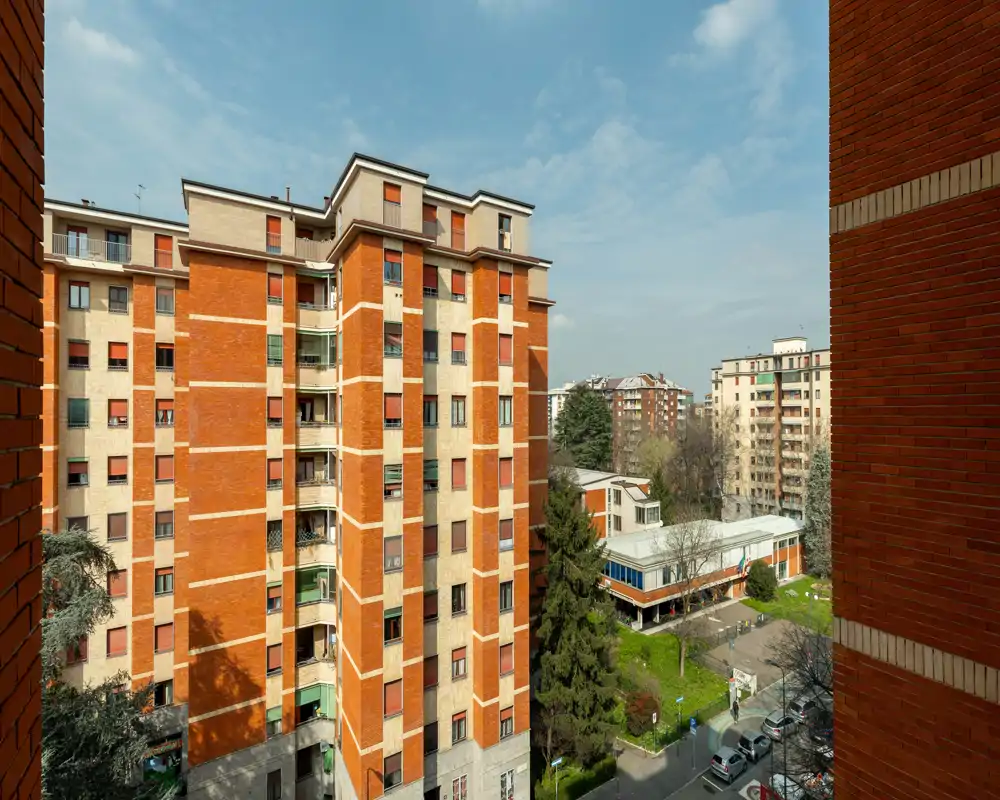 Vue extérieure lumineuse depuis une fenêtre montrant des immeubles en brique, une petite cour verte et la rue en contrebas — idéale pour présenter l'environnement urbain et l'exposition à la lumière.