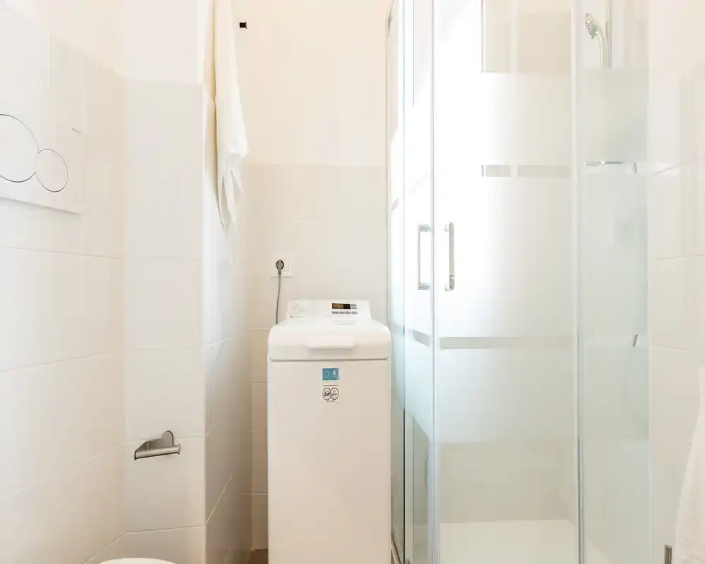 Salle de bains compacte et bien éclairée avec cabine de douche en verre et machine à laver à chargement par le dessus ; carrelage blanc et décoration minimale pour un style propre et moderne.