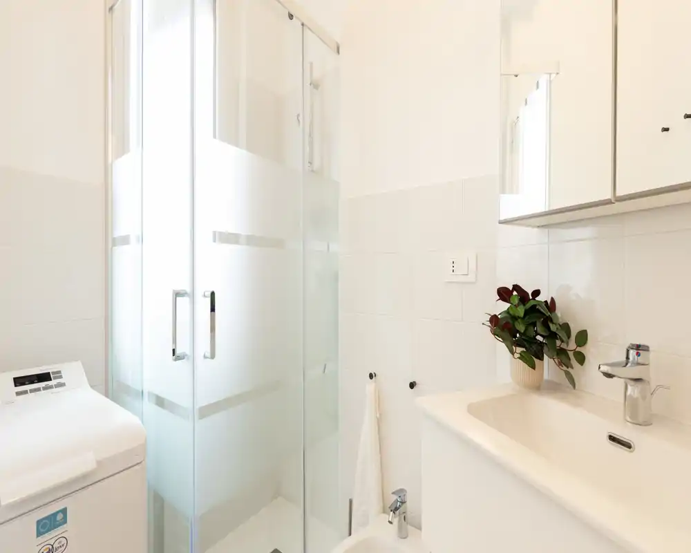 Salle de bains moderne et lumineuse avec douche vitrée, lavabo avec meuble et lave-linge à ouverture par le dessus. Carrelage blanc et lumière naturelle pour un espace propre et fonctionnel.