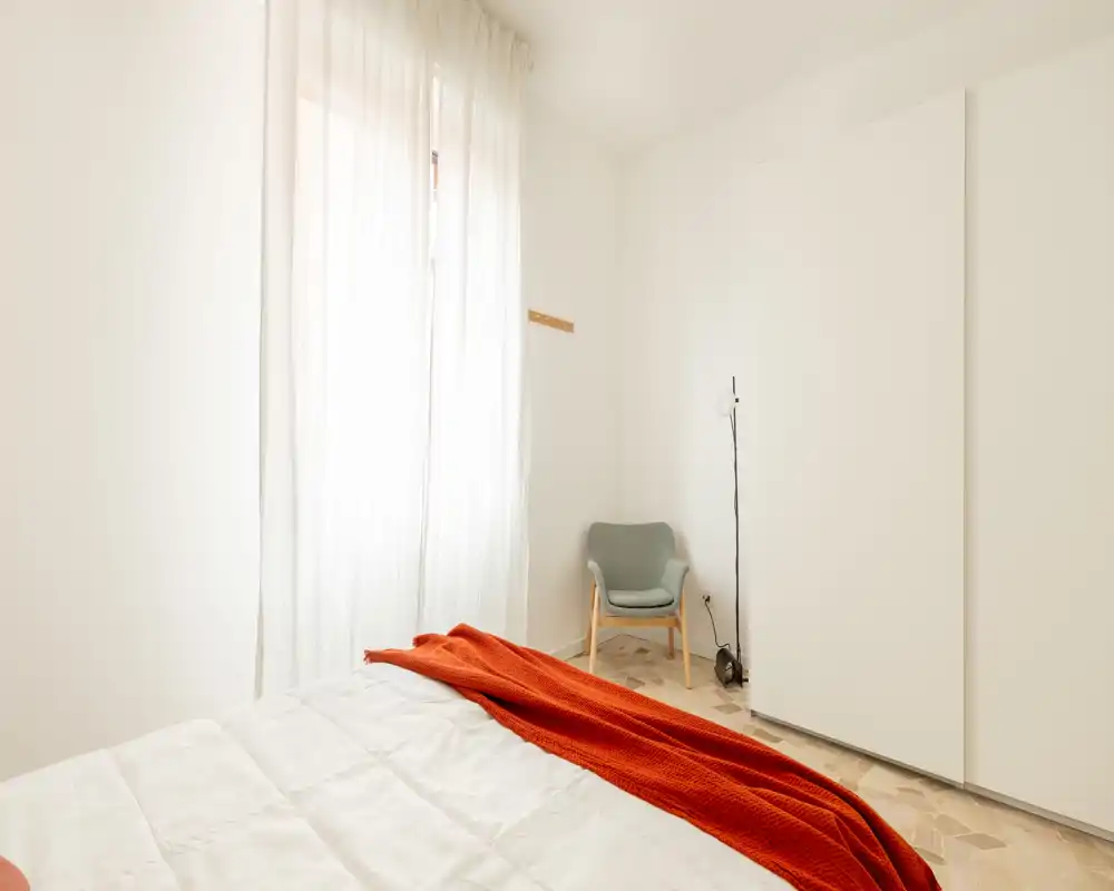 Camera da letto luminosa e minimalista con letto ben rifatto e un caldo plaid arancione, una poltroncina nell'angolo e un armadio bianco incassato — perfetta per un affitto moderno e tranquillo.