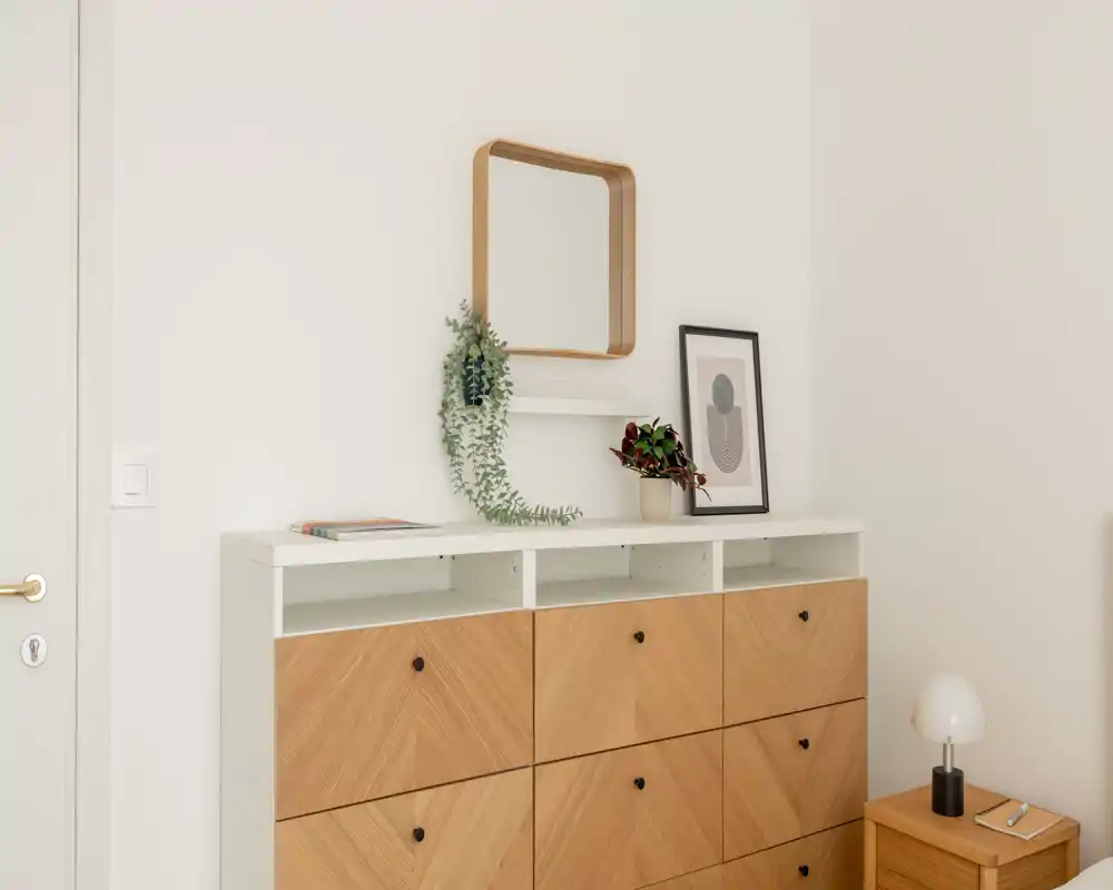 Ingresso luminoso e minimale con una consolle dai frontali in legno, uno specchio decorativo e una piccola lampada — angolo d'ingresso ordinato e accogliente.