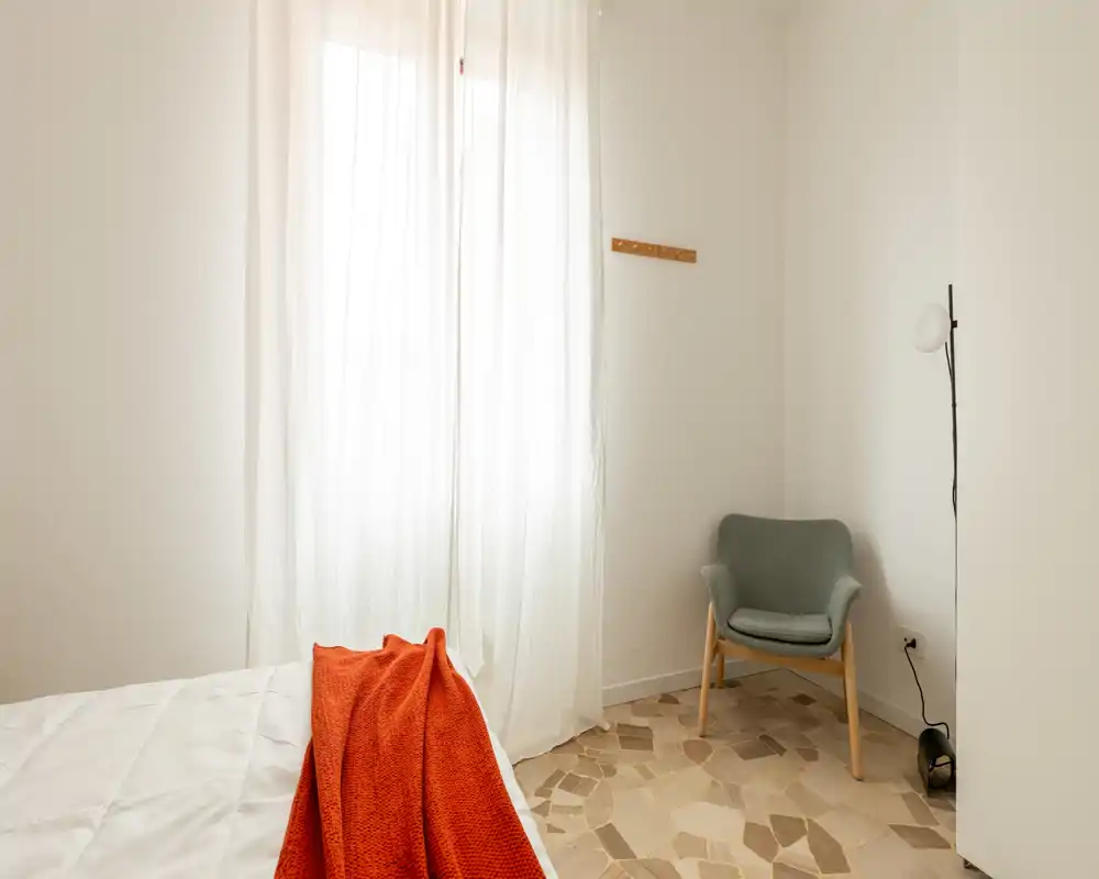 Angolo di camera luminoso e minimalista con letto parzialmente visibile, lenzuola bianche e plaid arancione, accanto a una poltrona moderna e una lampada da terra — perfetto per annunci accoglienti e soleggiati.