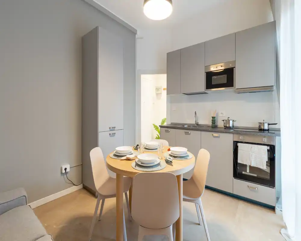 Cucina moderna con angolo pranzo: tavolo rotondo apparecchiato per quattro, mobili grigio chiaro, forno e microonde incassati e luce naturale soffusa da una finestra con tenda.