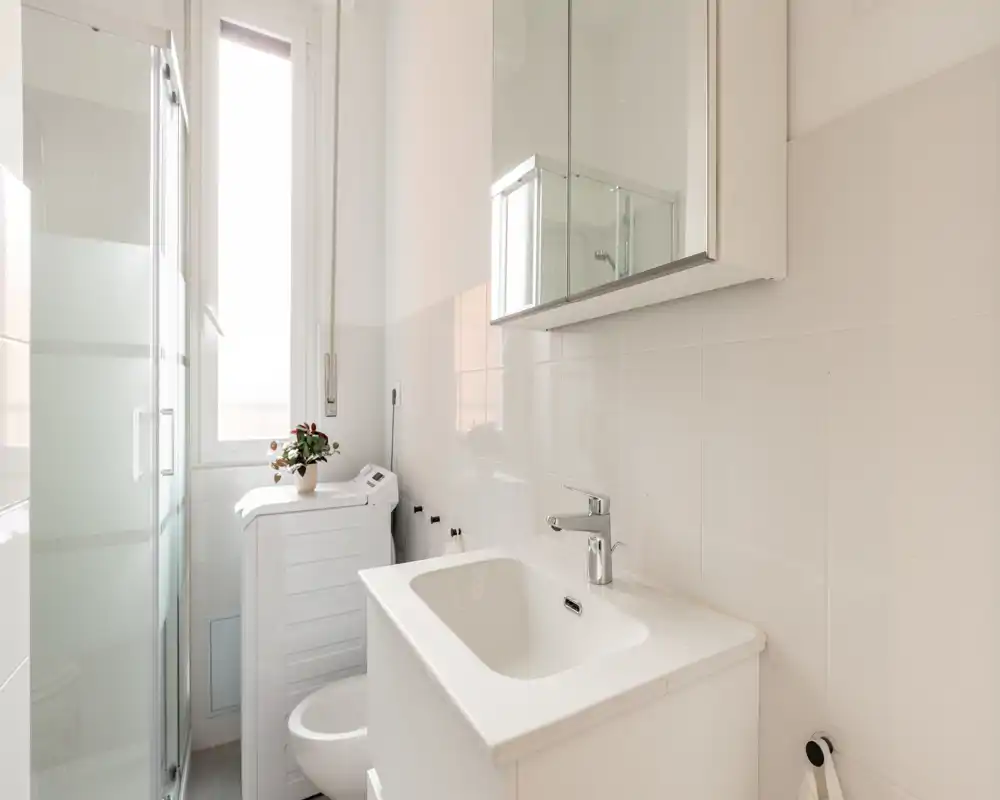 Bagno moderno e luminoso con lavabo, specchiera, box doccia e finestra alta che fornisce luce naturale; design compatto e pulito ideale per appartamenti in città.