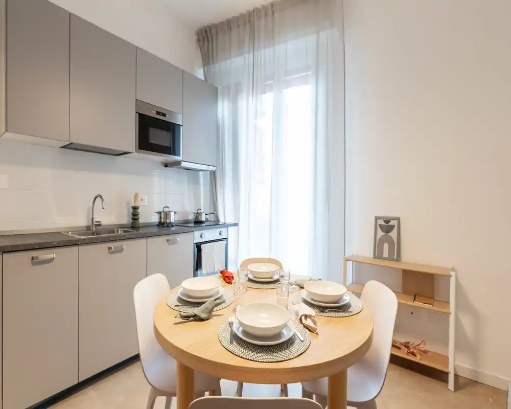 Angolo cucina e pranzo moderno con tavolo rotondo in legno apparecchiato per quattro, eleganti mobili grigi e luce naturale morbida da una grande finestra — uno spazio luminoso e dal design curato ideale per i pasti.