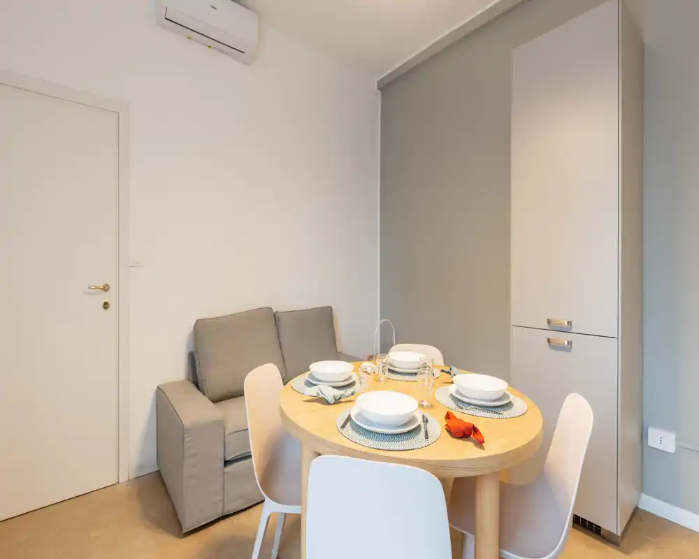 Accogliente angolo pranzo moderno con un tavolo rotondo in legno per quattro persone, sedute grigie e una dispensa alta — luminoso, essenziale e pronto per i pasti.