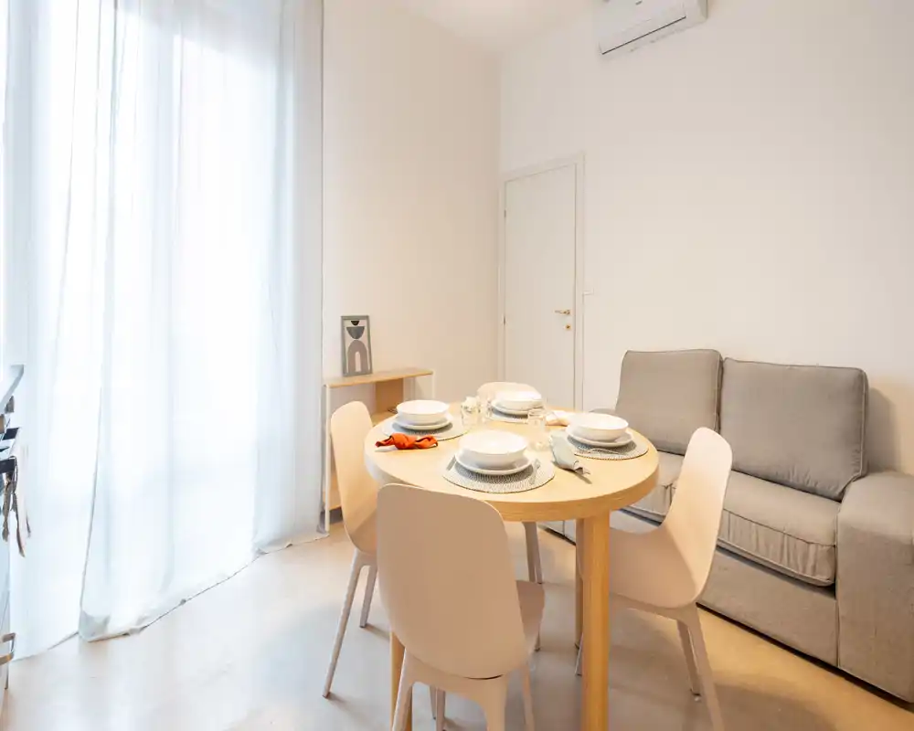 Zona pranzo luminosa e moderna con tavolo rotondo in legno apparecchiato per quattro, sedie morbide e un divano grigio compatto — perfetta per pasti informali in uno spazio minimal e accogliente.