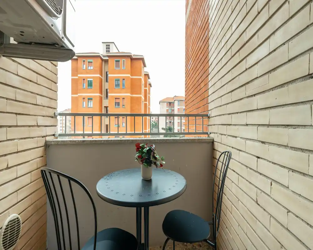 Piccolo balcone urbano con tavolino rotondo in metallo, due sedie e una pianta in vaso, con vista sui palazzi vicini.