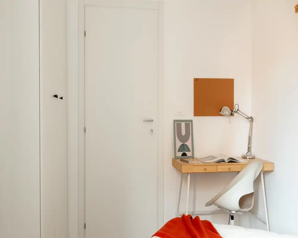 Angolo camera da letto luminoso e minimalista con una piccola scrivania in legno, lampada da lavoro e un lembo di letto con una coperta arancione — arredamento pulito e neutro ideale per un viaggiatore singolo.