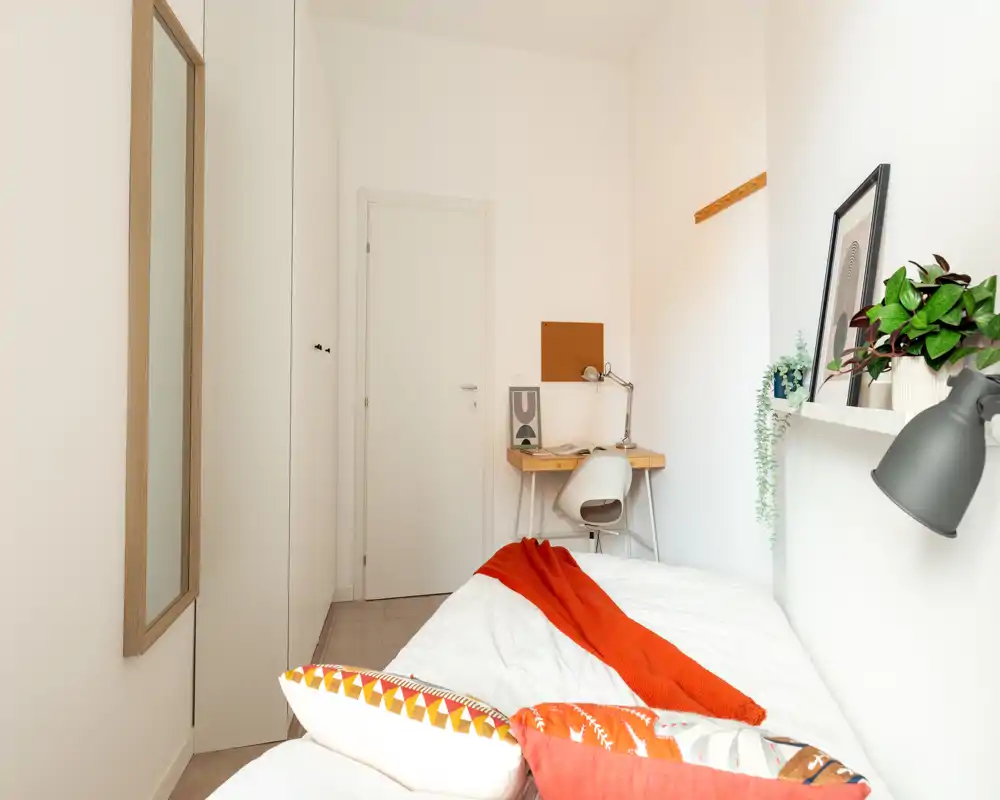 Camera da letto piccola e luminosa con letto singolo in lenzuola bianche e coperta arancione vivace; scrivania compatta con lampada vicino alla porta e piante e quadri che rendono l'ambiente accogliente.