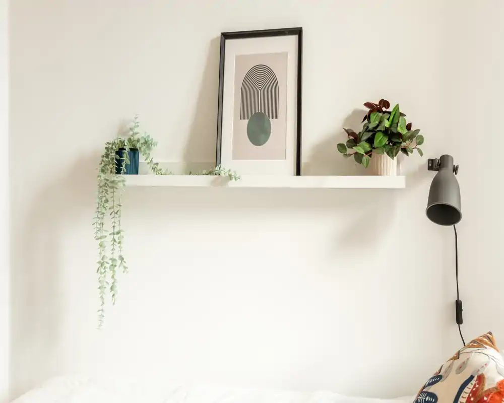 Angolo di camera da letto luminoso e minimal con mensola bianca, piante in vaso, quadro incorniciato e lampada da parete sopra il letto ordinato.