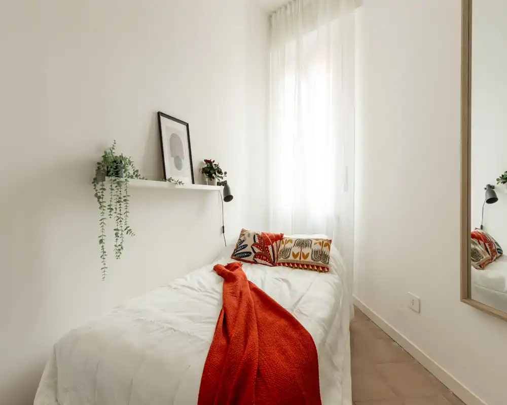 Camera singola luminosa e accogliente con letto bianco ben rifatto, cuscini decorativi colorati e una coperta arancione. La luce naturale attraversa tende leggere e una mensola con piante aggiunge un tocco minimal e fresco.