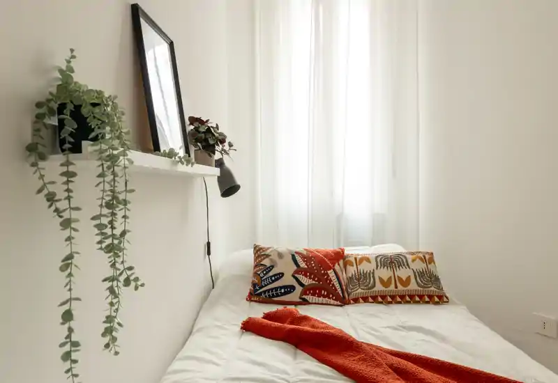 Camera da letto luminosa e minimale con cuscini decorativi e un caldo plaid arancione su un letto ben rifatto davanti a una finestra con tenda leggera.