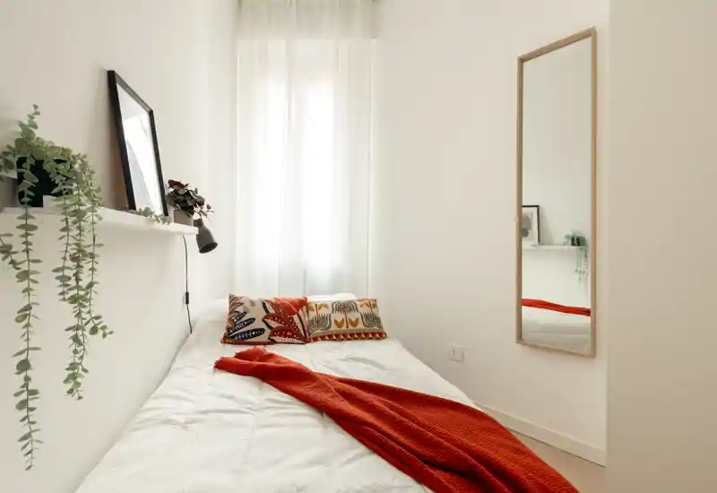 Camera da letto luminosa e minimalista con un letto singolo ben rifatto, cuscini decorativi e una calda coperta rossa. La luce naturale filtra dalle tende leggere e uno specchio a figura intera crea profondità.