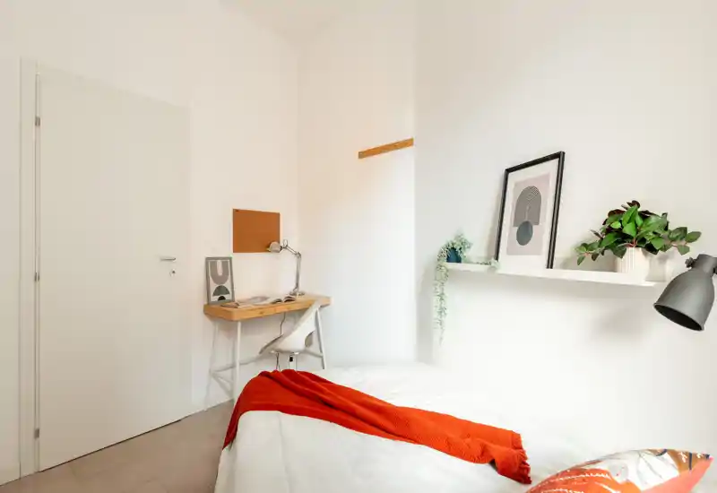 Camera da letto luminosa e minimal con letto ben rifatto e un plaid rosso vivace, una scrivania compatta nell’angolo e mensola con quadro e piante — perfetta per annunci per una persona.