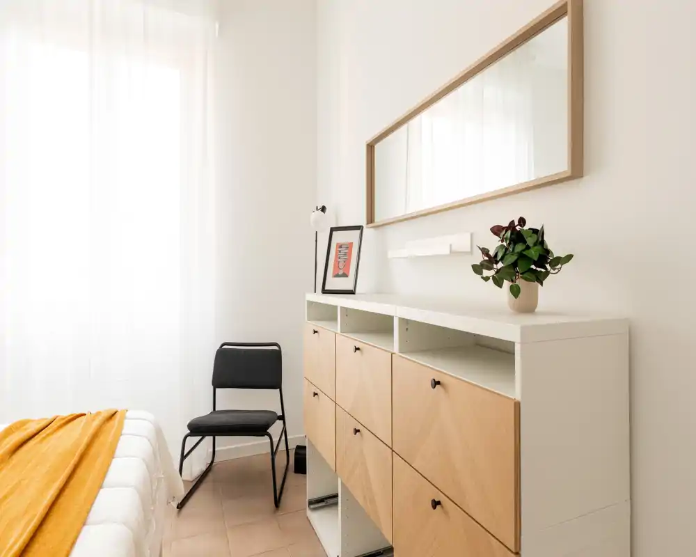 Camera da letto luminosa e minimale con letto ben rifatto e una coperta color senape, una sedia semplice, una cassettiera bassa con frontali in legno e uno specchio a parete — arredamento in stile scandinavo.