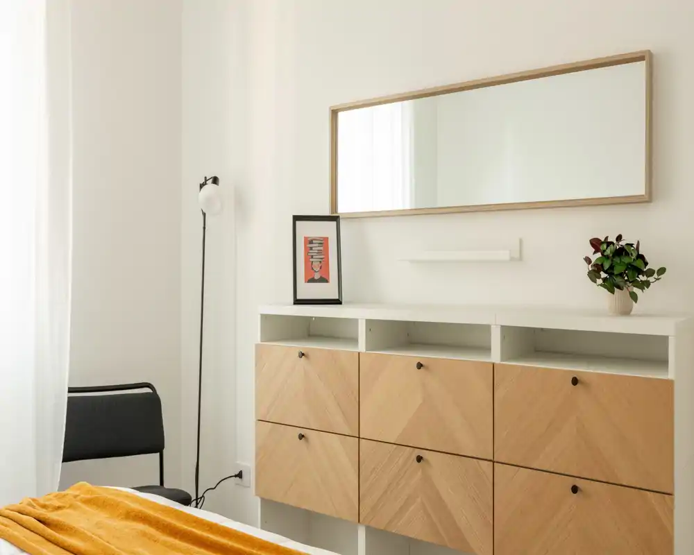 Angolo di camera luminoso e minimal con un mobile contenitore in legno, grande specchio a parete, lampada da terra e parte del letto con biancheria calda.