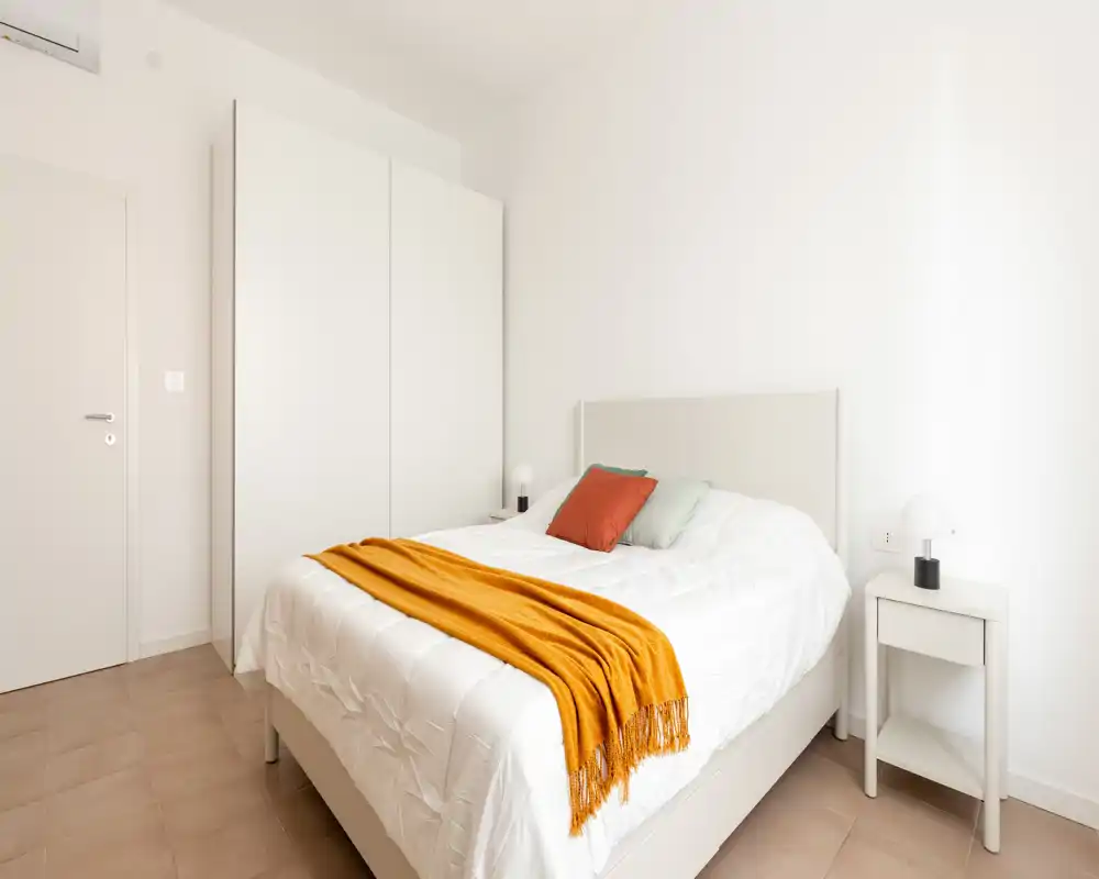 Camera da letto luminosa e minimal con letto matrimoniale bianco, plaid accogliente e comodini; toni neutri e linee pulite creano un'atmosfera moderna e tranquilla.