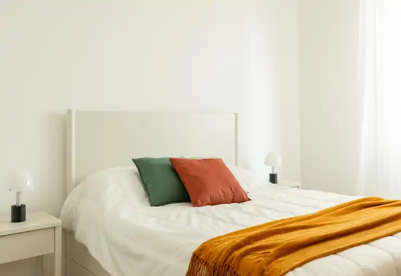 Camera da letto luminosa e minimal con testiera bianca, due comodini e cuscini e plaid dai toni caldi che creano un’atmosfera accogliente e moderna.