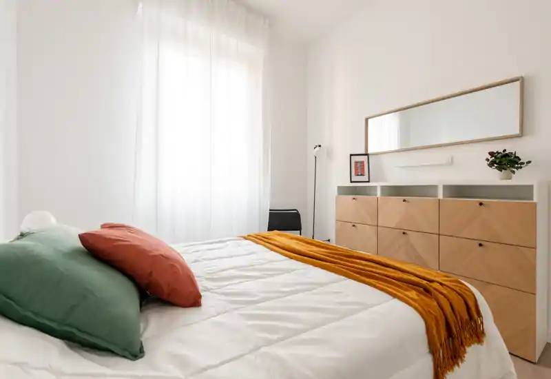 Camera da letto luminosa e minimale con letto ben rifatto, cuscini colorati e una coperta calda; un ampio comò con specchio e luce naturale morbida dalla finestra creano un ambiente arioso e accogliente.