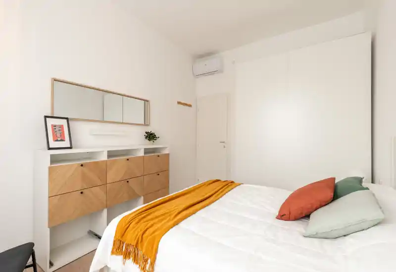 Camera da letto luminosa e moderna con letto ben rifatto, sciarpa e cuscini colorati, comò in legno e ampia armadiatura bianca a muro; stile minimal pulito, perfetto per annunci.