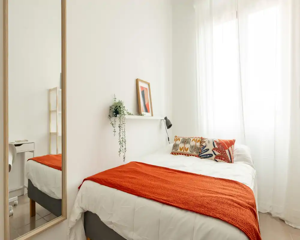 Camera da letto luminosa e accogliente con letto ben rifatto, cuscini decorativi e un plaid arancione; la luce naturale morbida dalla finestra rende l'ambiente caldo e invitante.