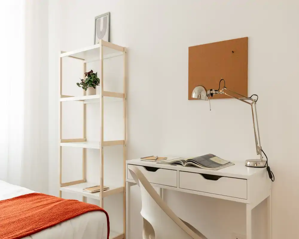 Angolo camera luminoso e minimal con scrivania bianca e scaffale accanto a un letto ordinato con plaids arancione; la luce naturale crea un’atmosfera pulita e accogliente.