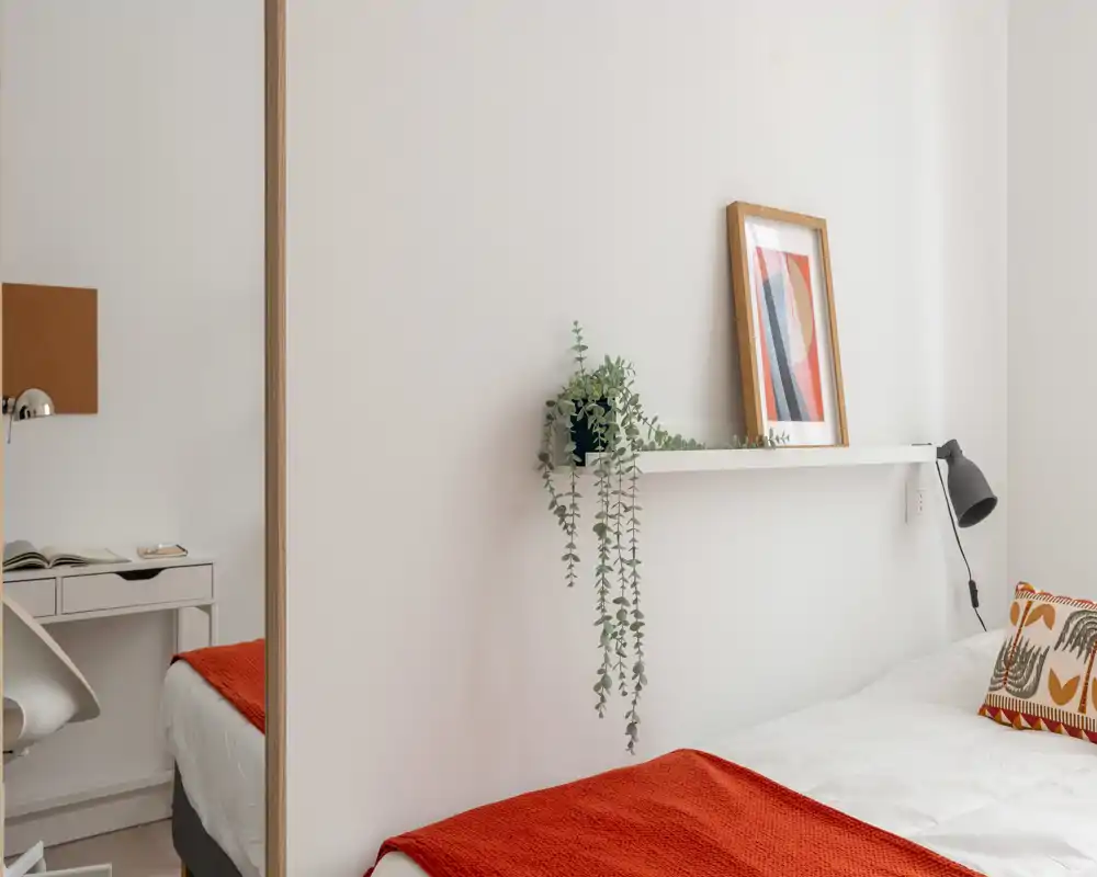 Camera da letto luminosa e minimal con letto bianco, coperta arancio e cuscino decorativo; una mensola con pianta e cornice aggiunge calore.
