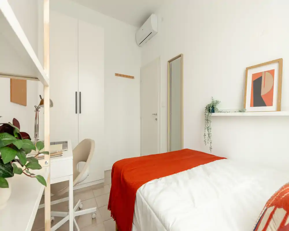 Camera da letto luminosa e compatta con armadio bianco, scrivania e letto ben rifatto con un caldo plaid rosso. Arredo minimal con piante che creano un’atmosfera accogliente, ideale per una persona.