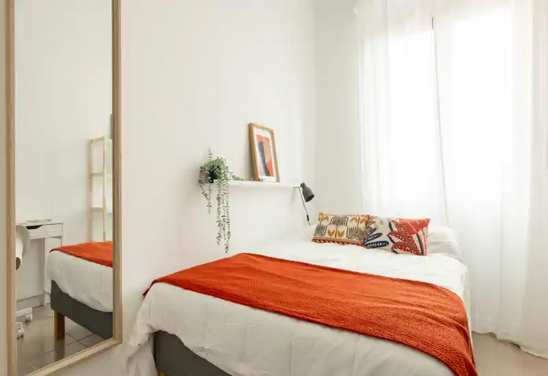 Camera da letto luminosa e accogliente con letto ben rifatto, cuscini decorativi e un plaid arancione; la luce naturale morbida dalla finestra rende l'ambiente caldo e invitante.