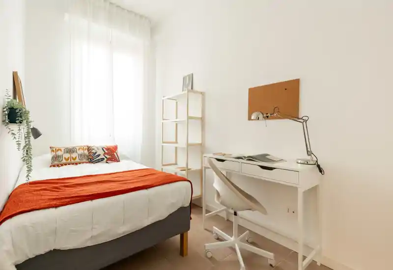 Camera da letto luminosa e compatta con un comodo letto matrimoniale, cuscini decorativi e un plaid arancione. Una piccola scrivania bianca con lampada moderna e scaffalatura crea un pratico angolo lavoro accanto alla finestra con tende leggere.